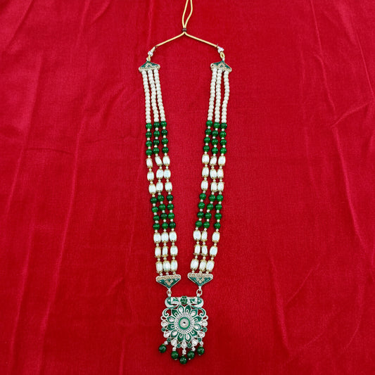 A traditional Indian necklace featuring white pearl strands, green beads, and a large green and silver floral pendant, displayed on a red fabric background.
